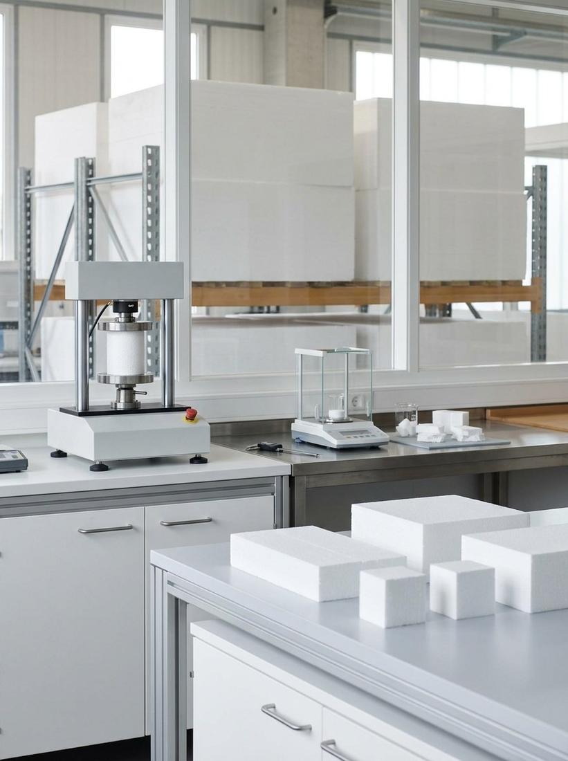 Insutech's in-house materials lab — a compression testing machine, a precision balance and cut EPS specimens on the bench, with large white expanded-polystyrene blocks stacked on racks behind